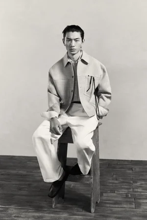 A seated male model wearing a light-patterned jacket and white trousers poses indoors against a plain wall for a Zara fashion project