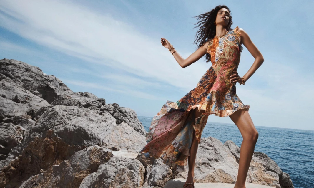 Woman in flowing floral dress poses on rocky coast with ocean backdrop under blue sky, fashion editorial image in outdoor setting.