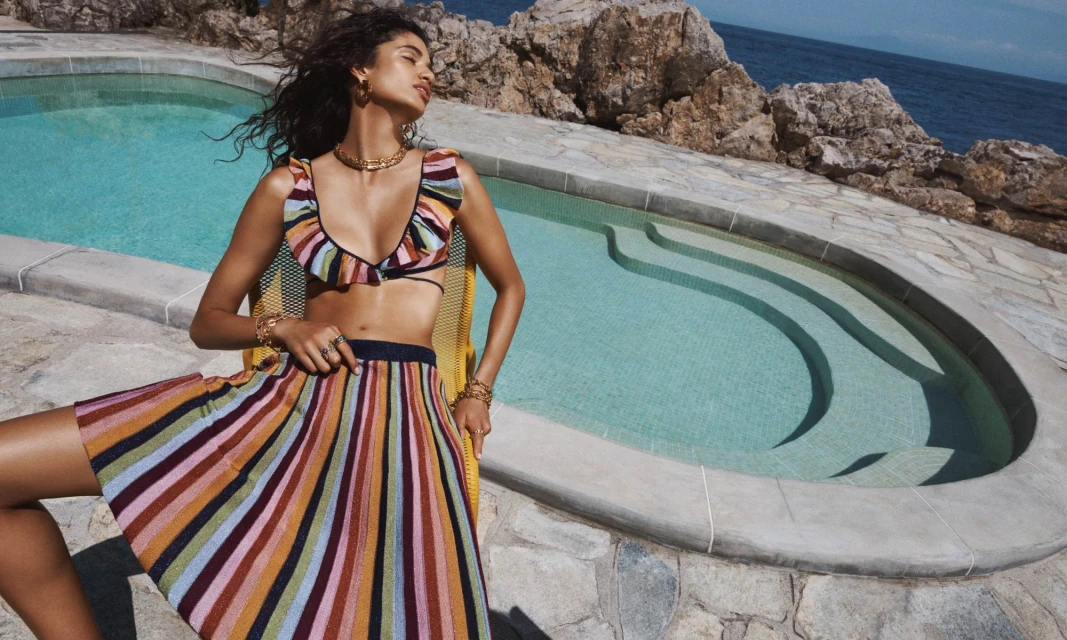 Woman in striped bikini top and skirt poses by a pool with blue water and rocky cliffs in the background on a sunny day.
