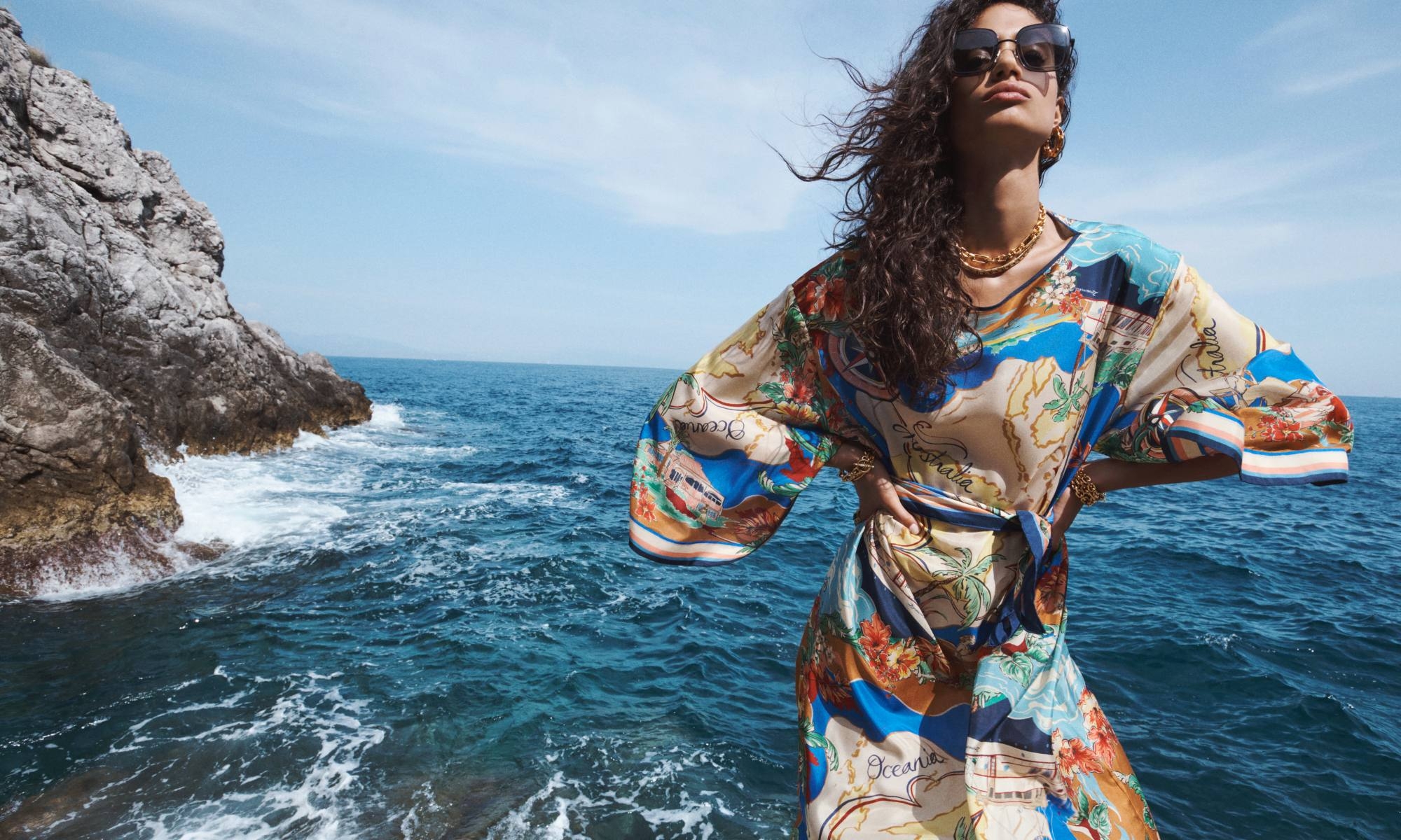 A woman in a patterned dress poses on rocks by the ocean under a blue sky with some clouds, wearing sunglasses and jewelry.