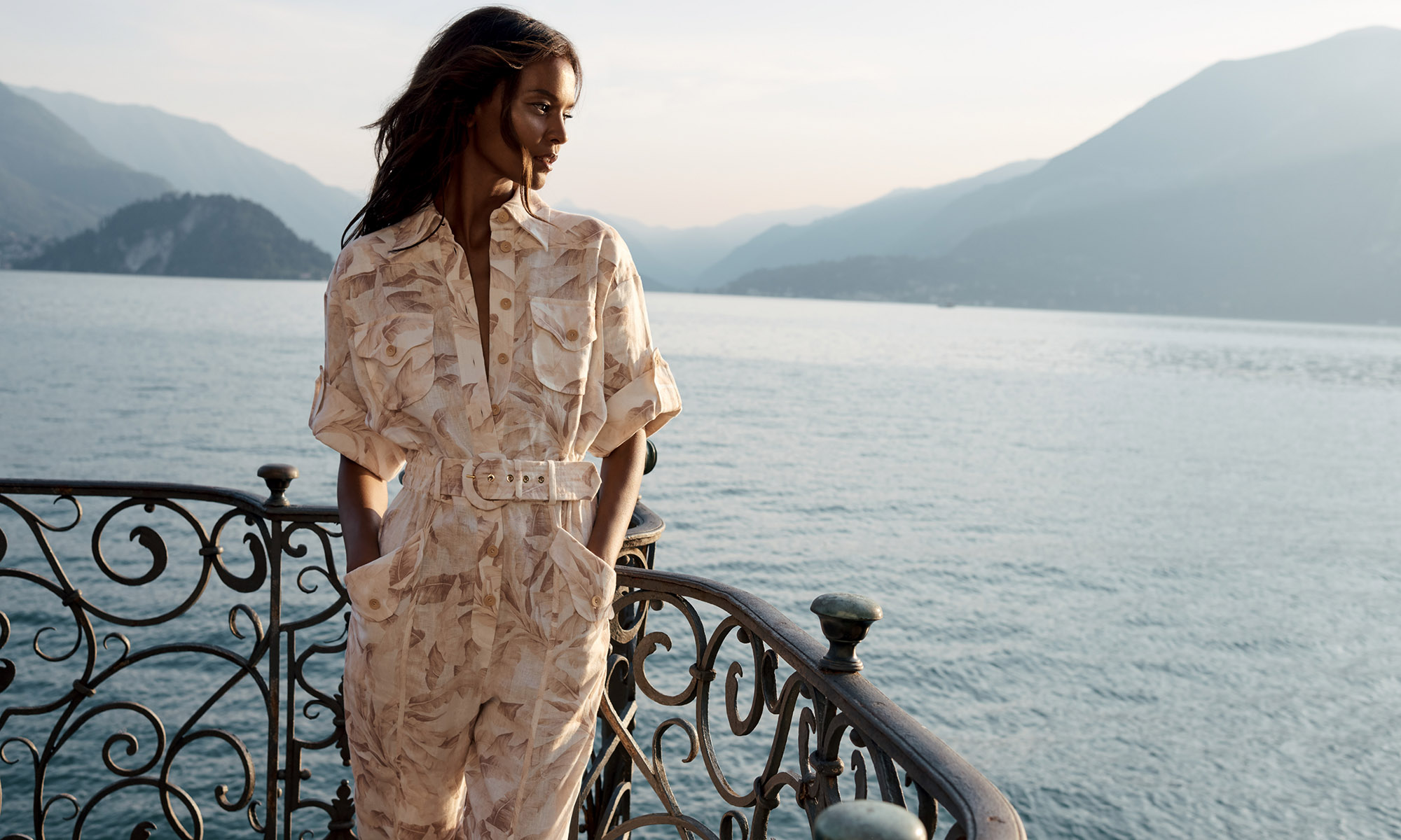 Adult female stands on balcony wearing jumpsuit overlooking Lake Como with mountains in the background on a sunny day.