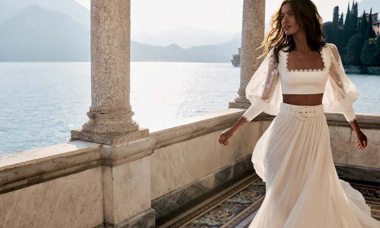 Brunette fashion model in white crop top and flowing skirt stands on a stone terrace with a lake and mountains in the background.