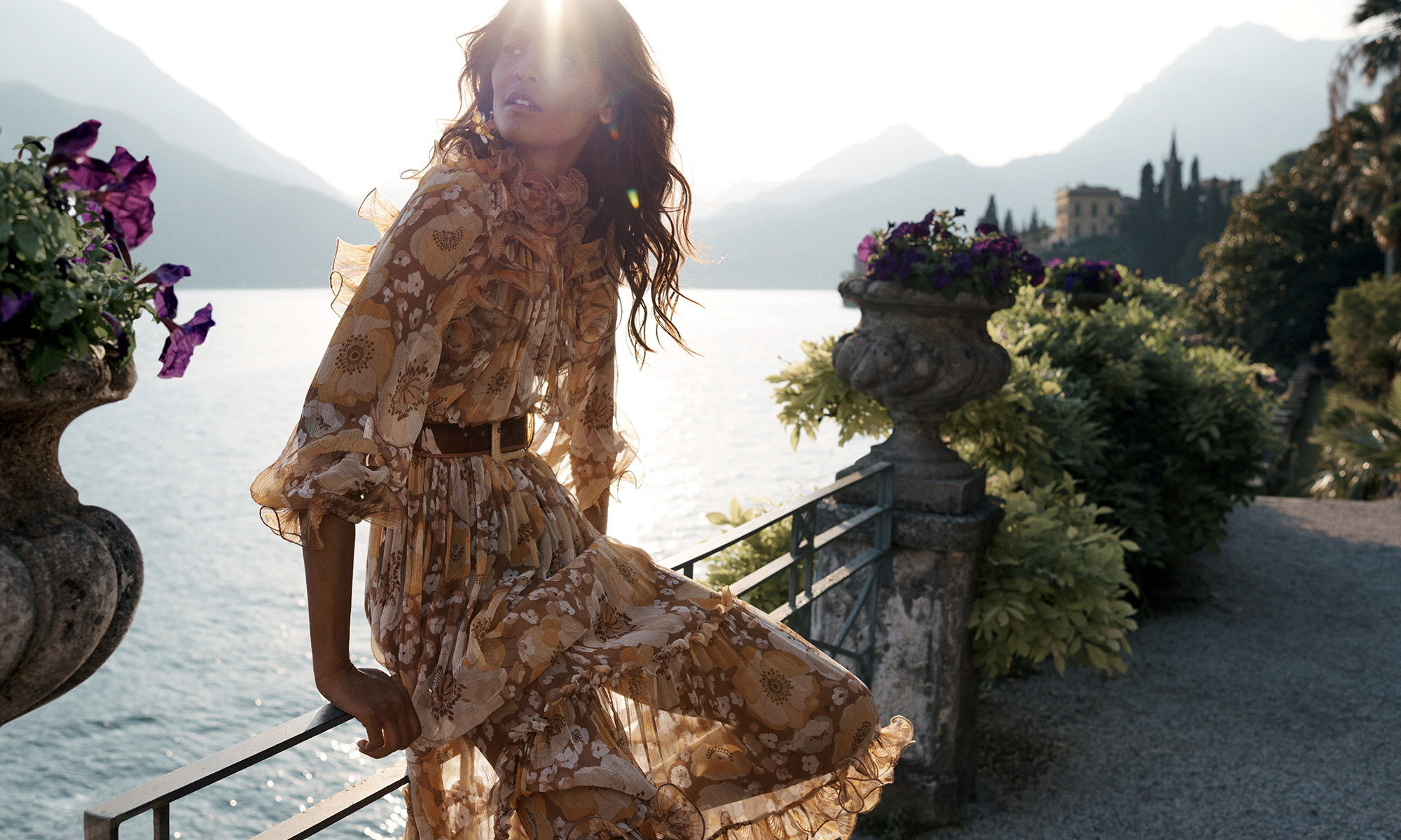 A woman in a patterned dress poses on a stone terrace overlooking a lake and mountains in a fashion shot, bathed in sunlight.