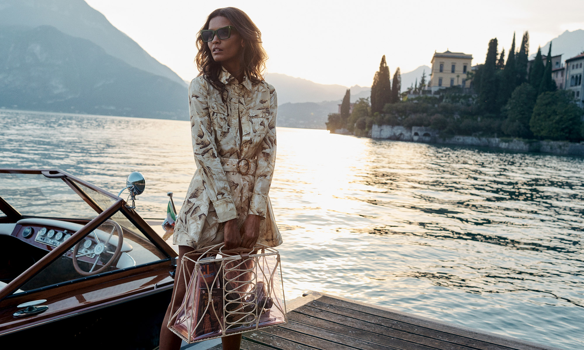 Woman in patterned dress with handbag on boat, set against scenic lake and mountains, captured in soft, natural light.