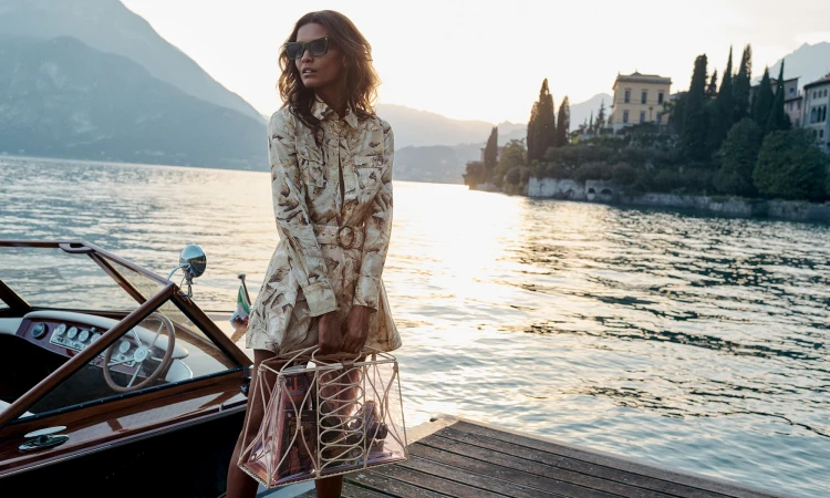 Woman in patterned dress with handbag on boat, set against scenic lake and mountains, captured in soft, natural light.