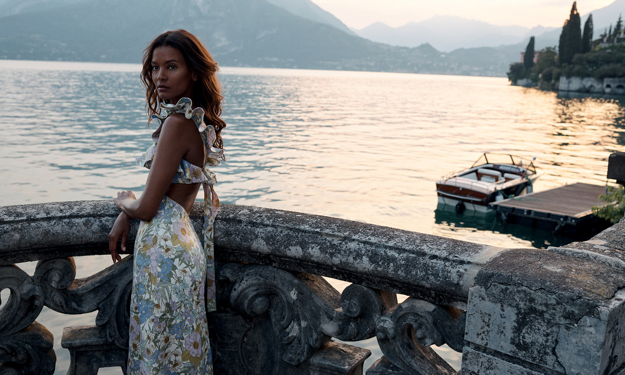 A woman in a floral dress stands by a stone railing overlooking a lake with mountains in the background in a picturesque outdoor setting.