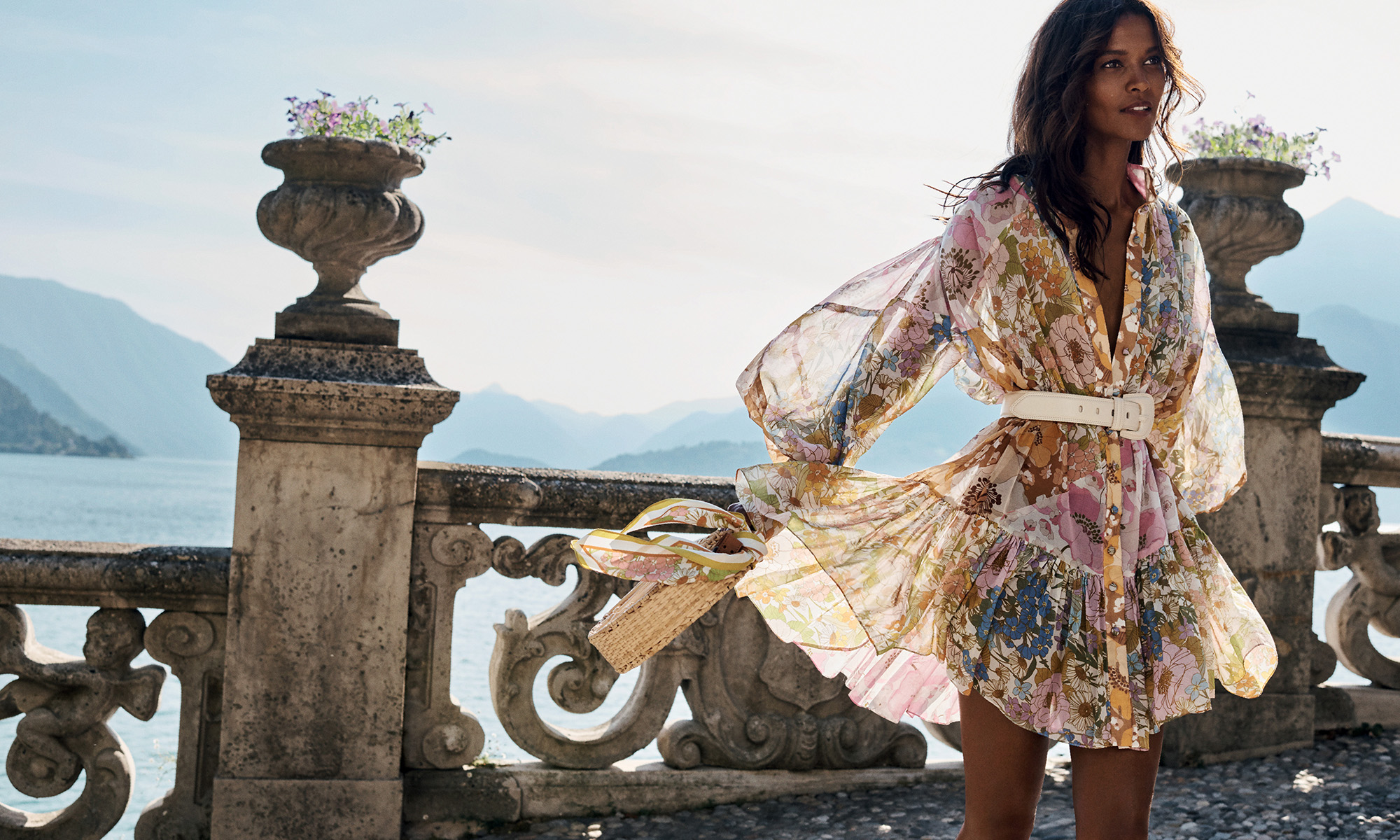 Elegant woman in a floral dress poses on a stone terrace with a purse, overlooking a scenic lake and mountain vista on a sunny day.