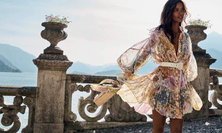 Elegant woman in a floral dress poses on a stone terrace with a purse, overlooking a scenic lake and mountain vista on a sunny day.