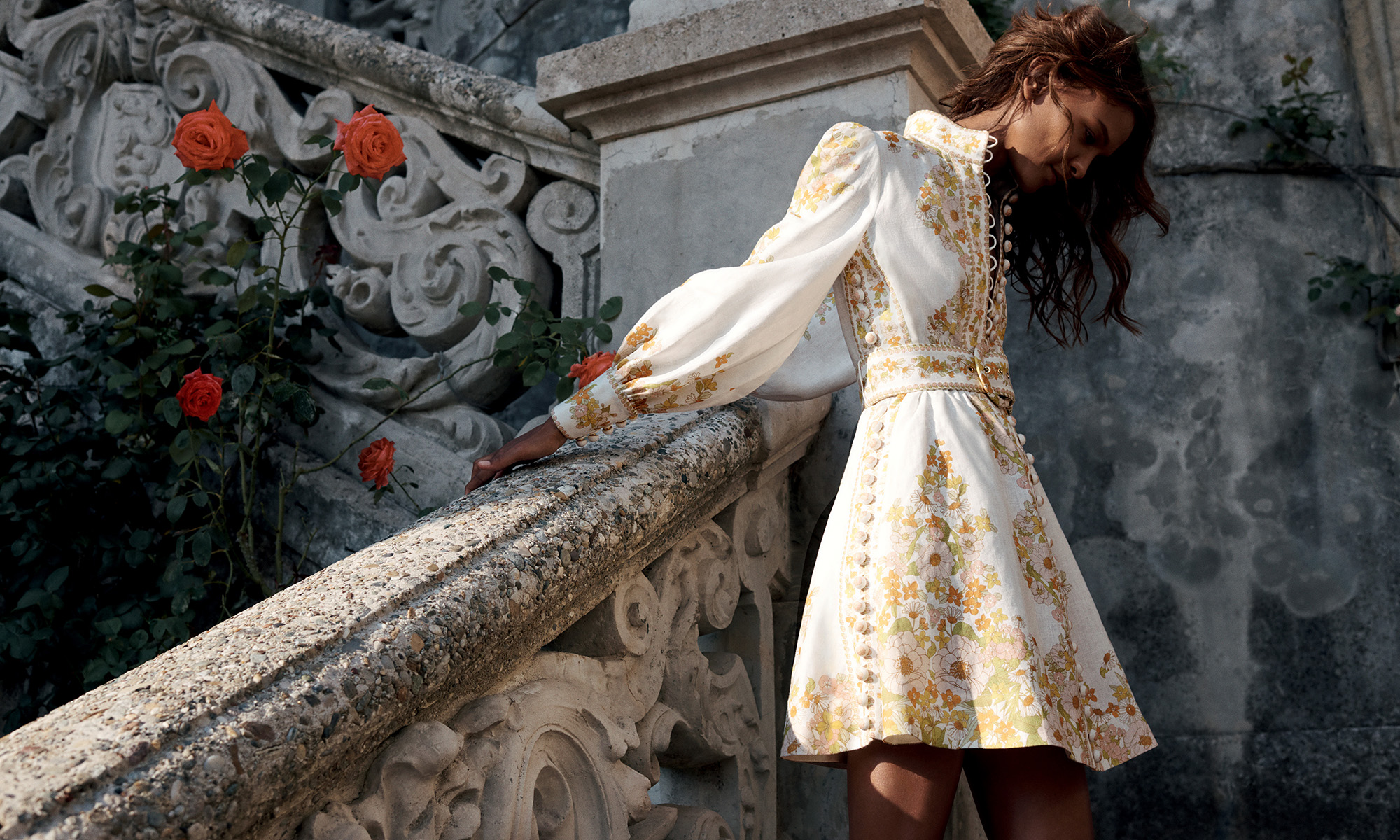 Adult female in a floral-patterned dress poses near a stone staircase and rose bushes in a full shot with a low eye-level angle.