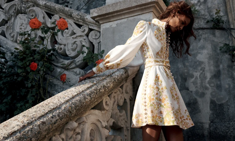 Adult female in a floral-patterned dress poses near a stone staircase and rose bushes in a full shot with a low eye-level angle.