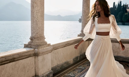 Model in white crop top and belted maxi skirt walking on a stone balcony overlooking a lake.