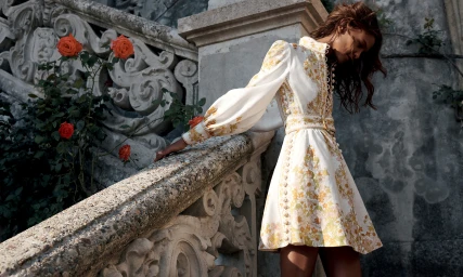 Model wearing a white floral patterned dress leaning on an ornate stone staircase with orange roses.