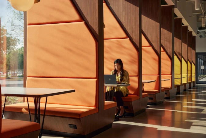 Woman works on a laptop in a modern office space with orange booth seating, wood dividers, and natural light, creating a productive atmosphere.