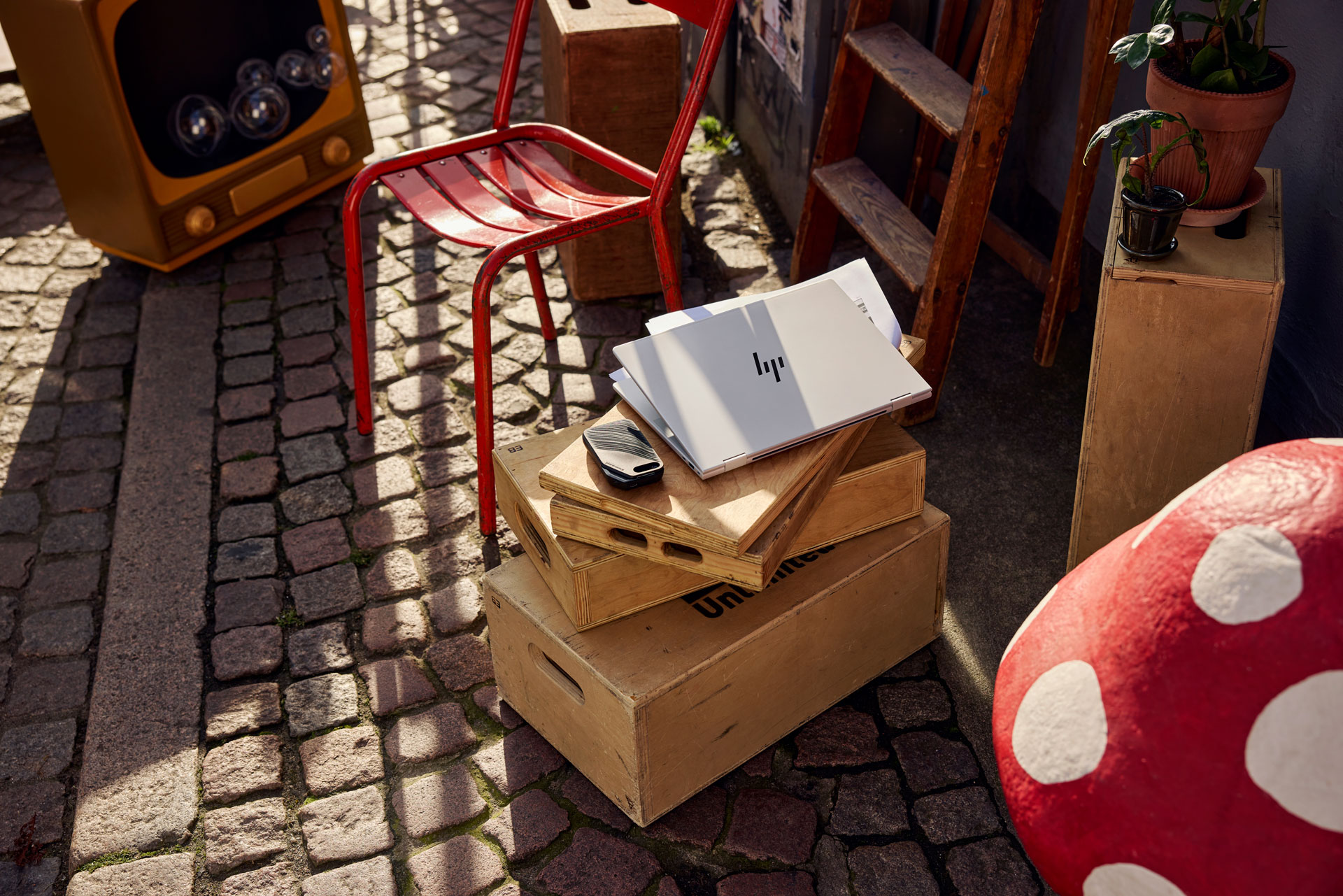 A rustic outdoor scene featuring a laptop on wooden boxes next to a red metal chair, a vintage TV, and a whimsical mushroom stool.