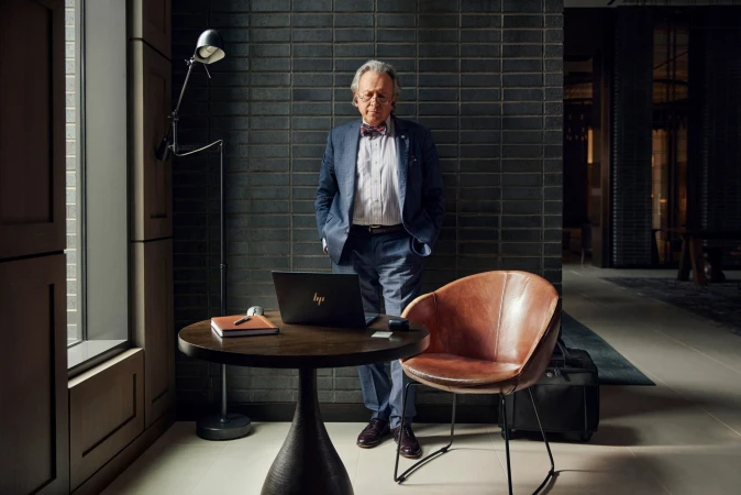 Mature man in a suit poses next to a table with a laptop, notebook, and pen in a dimly lit room with a brick accent wall.