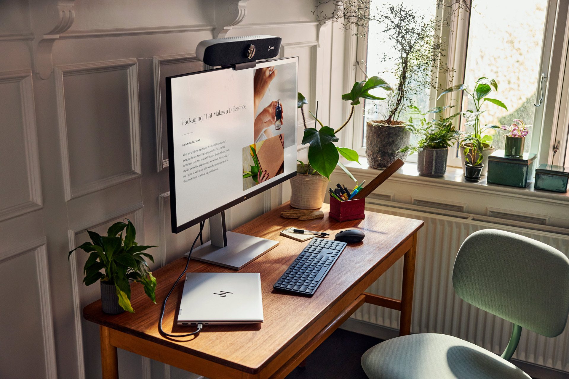 A modern home office setup featuring a wooden desk with a laptop, monitor, keyboard, mouse, plants, and natural light from a window.