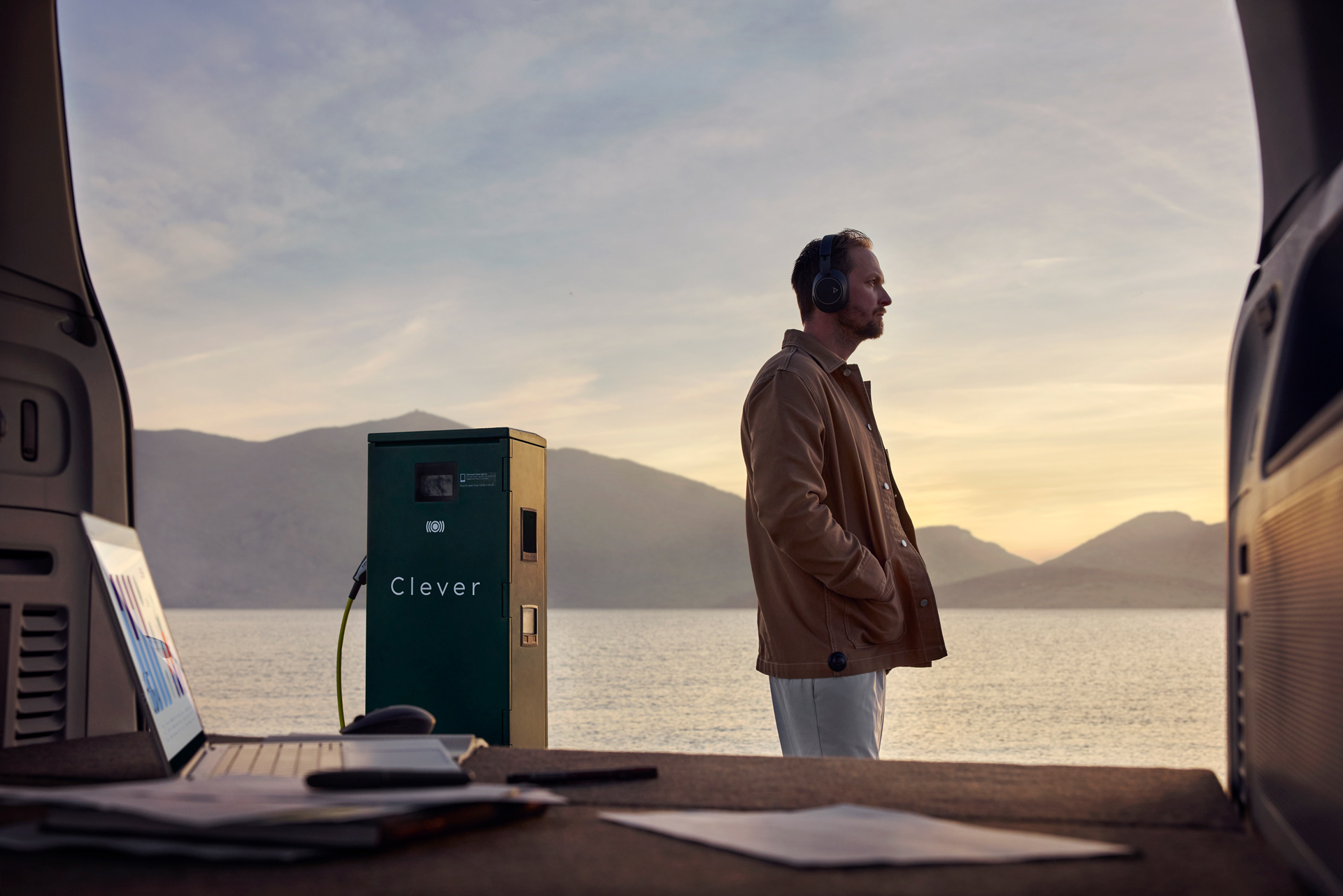 Man wearing headphones stands near an electric vehicle charging station by the water, viewed from inside vehicle trunk.