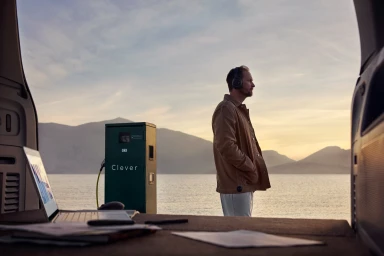 Man wearing headphones standing by open vehicle trunk facing a scenic lake and mountains at sunset