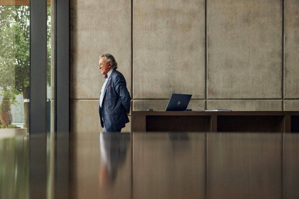 An elderly man in a blue suit stands in a modern interior, near a window with greenery outside, beside a laptop and papers on a desk.