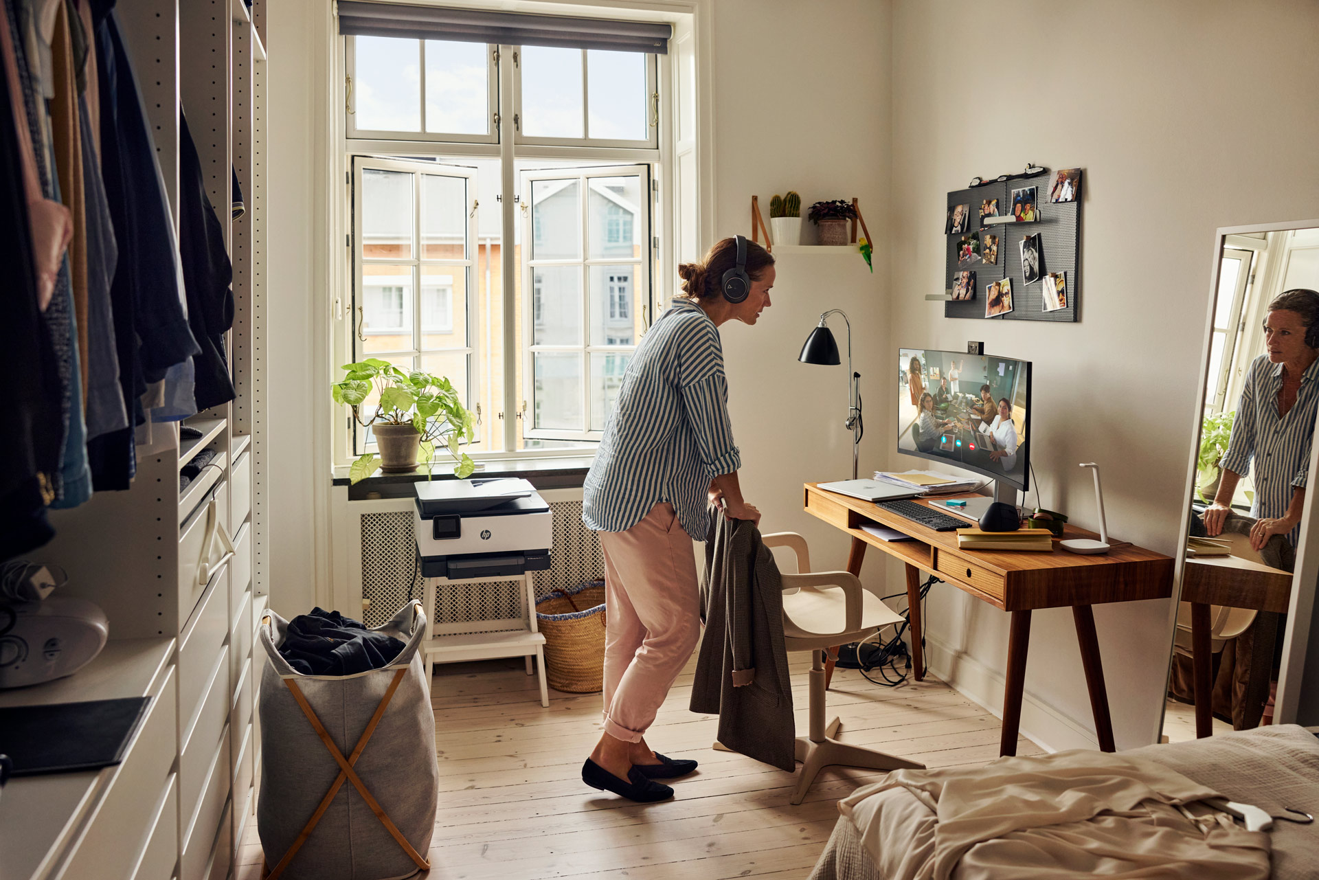 Woman with headphones stands in a bedroom with a computer desk and video call visible on the computer screen in natural daylight.