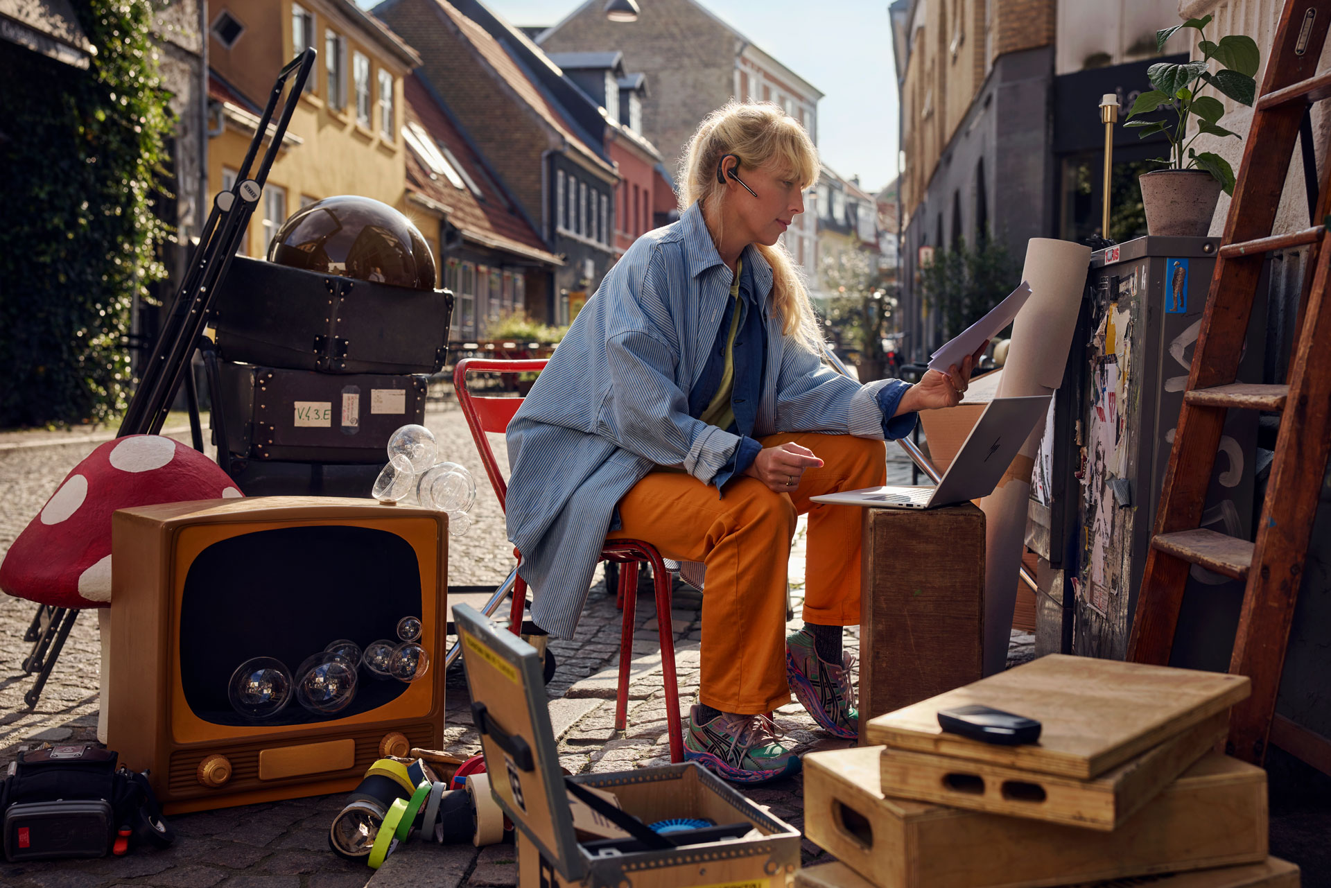 Woman with blonde hair works on a laptop in an outdoor alleyway. Eclectic collection of objects surrounds her, including boxes and TV.