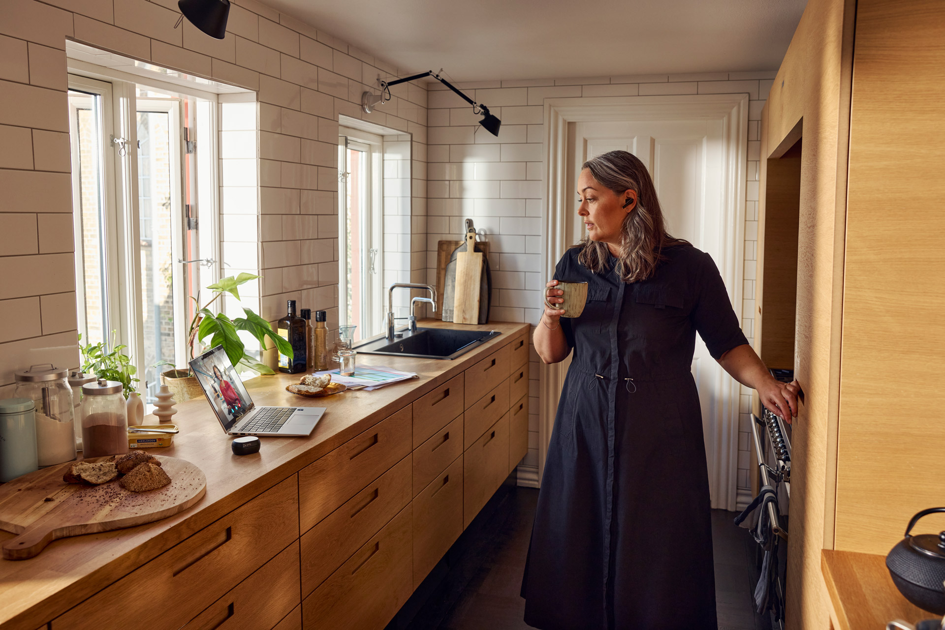 Adult woman with gray hair holds a mug while standing in a kitchen with a laptop and cooking utensils on the counter.