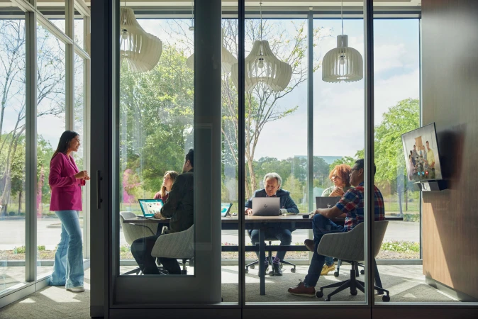 Modern office meeting in a glass-enclosed conference room with a woman standing and presenting to a diverse group around a table.