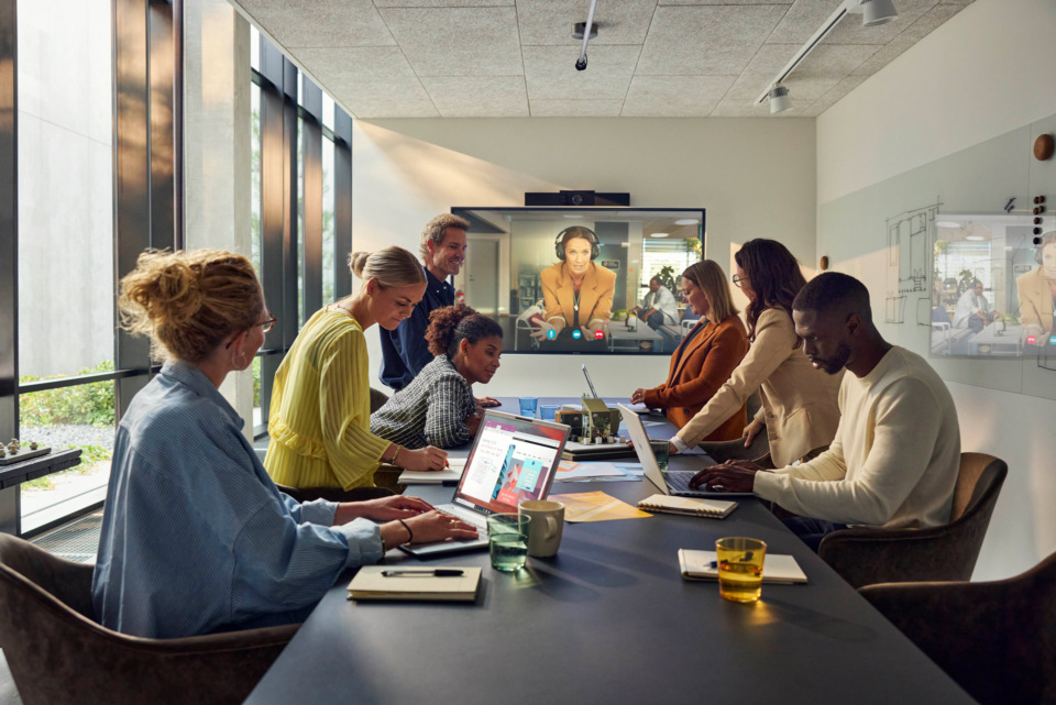 Group of professionals in a modern conference room using laptops during a video call, featuring a diverse team in a collaborative workspace.