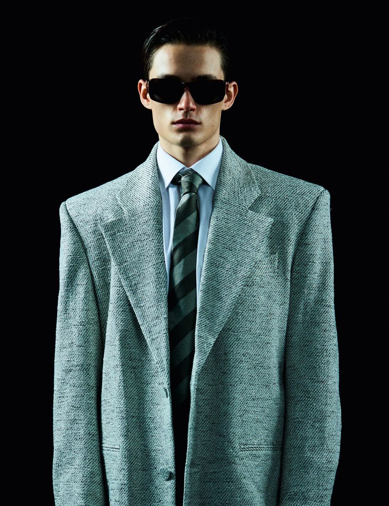 Portrait of a young adult male in a suit, wearing dark sunglasses, standing against a black backdrop in a studio setting.