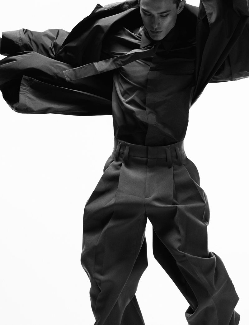 Monochrome fashion portrait of male model Alex Schlab in dark pleated trousers and dynamic windblown jacket