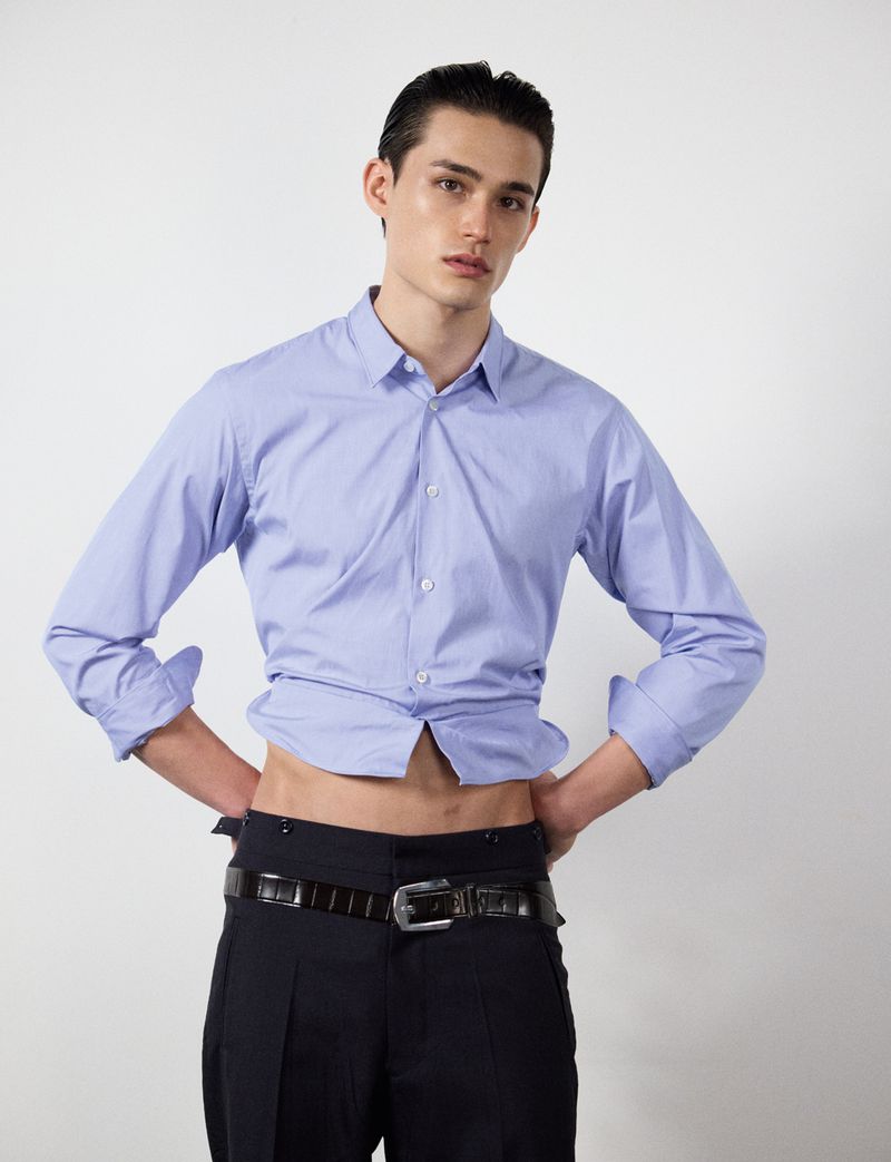 Model wearing a light blue buttoned shirt and dark trousers against a white seamless background for HERO Magazine