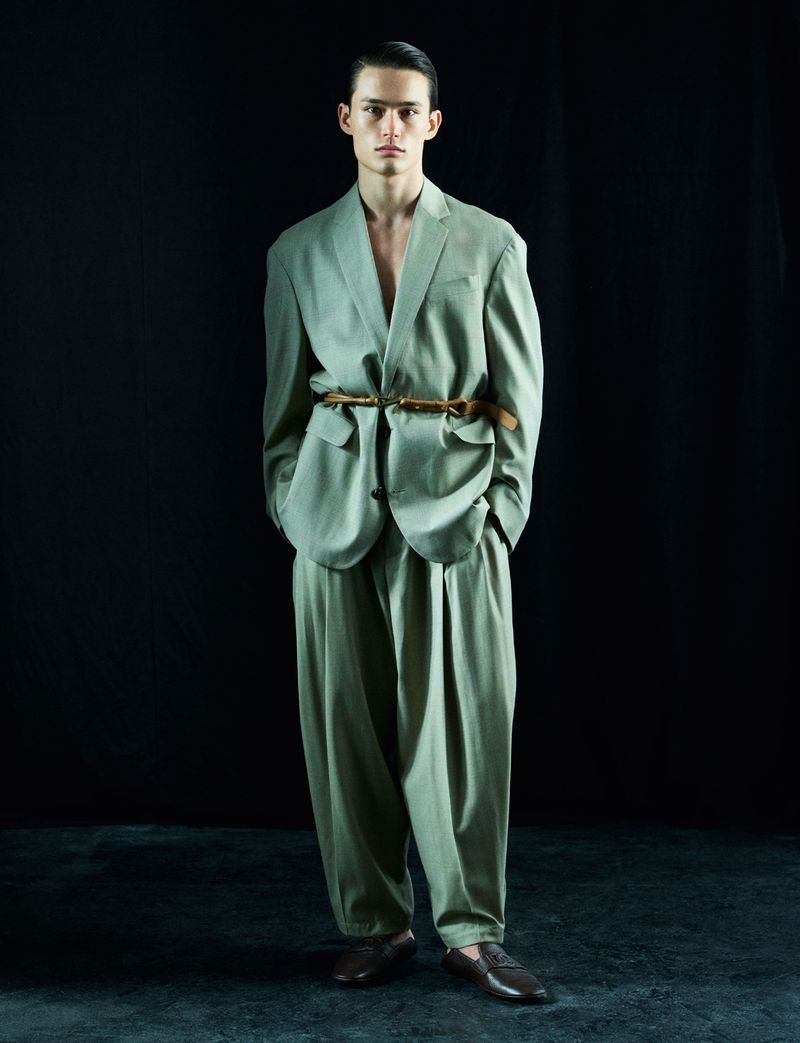A young adult male stands in a suit with a belt around the jacket, full shot, with a solid black background and grey flooring.