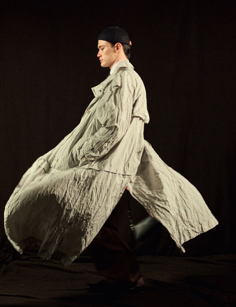 Young adult male fashion model showcases a flowing grey coat, dark pants and cap in a studio setting against a dark background.
