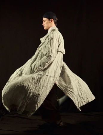 Young adult male fashion model showcases a flowing grey coat, dark pants and cap in a studio setting against a dark background.