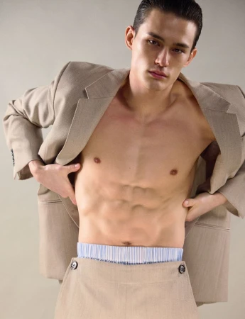 Fashion shot of a male model wearing a blazer and skirt, showcasing his abs against a neutral backdrop in a studio setting.