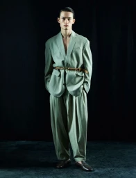 Model wearing a loose light green suit cinched with a thin belt against a dark background for HERO Magazine