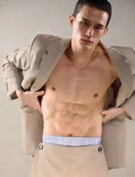 Shirtless male model wearing an open beige suit jacket and matching trousers with a striped waistband against a seamless grey backdrop