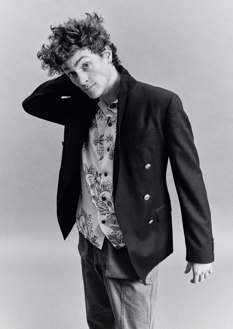 Black and white portrait of curly-haired man in blazer and patterned shirt, posing with hand behind neck against plain backdrop.