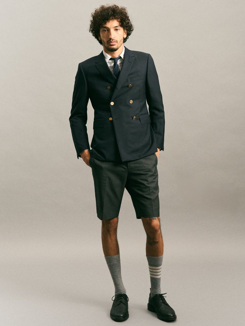 Full-length studio shot of a man in a navy blazer, dress shirt, tie, and grey shorts, accessorized with socks and dress shoes.