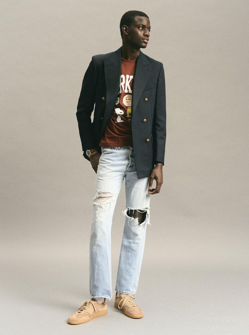 Dark-skinned male fashion model wears a black blazer, t-shirt with comic-strip graphic, and ripped jeans against a neutral background.
