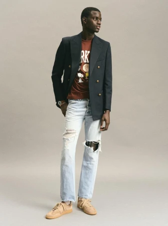 Dark-skinned male fashion model wears a black blazer, t-shirt with comic-strip graphic, and ripped jeans against a neutral background.