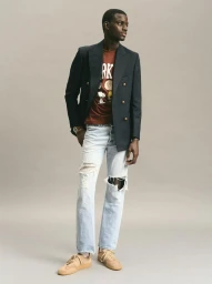Model wearing a navy blazer over a graphic t-shirt and distressed light-wash jeans against a neutral grey studio background