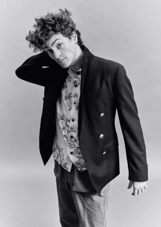 Monochrome portrait of a young man with curly hair wearing a dark double-breasted blazer and patterned shirt against a grey backdrop