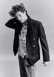 Monochrome portrait of a young man with curly hair wearing a dark double-breasted blazer and patterned shirt against a grey backdrop