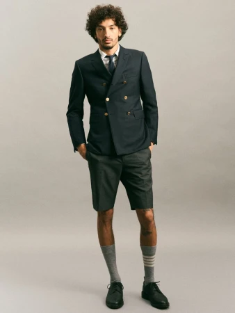 Man wearing a navy double-breasted blazer, dark shorts, and grey crew socks against a neutral grey studio background
