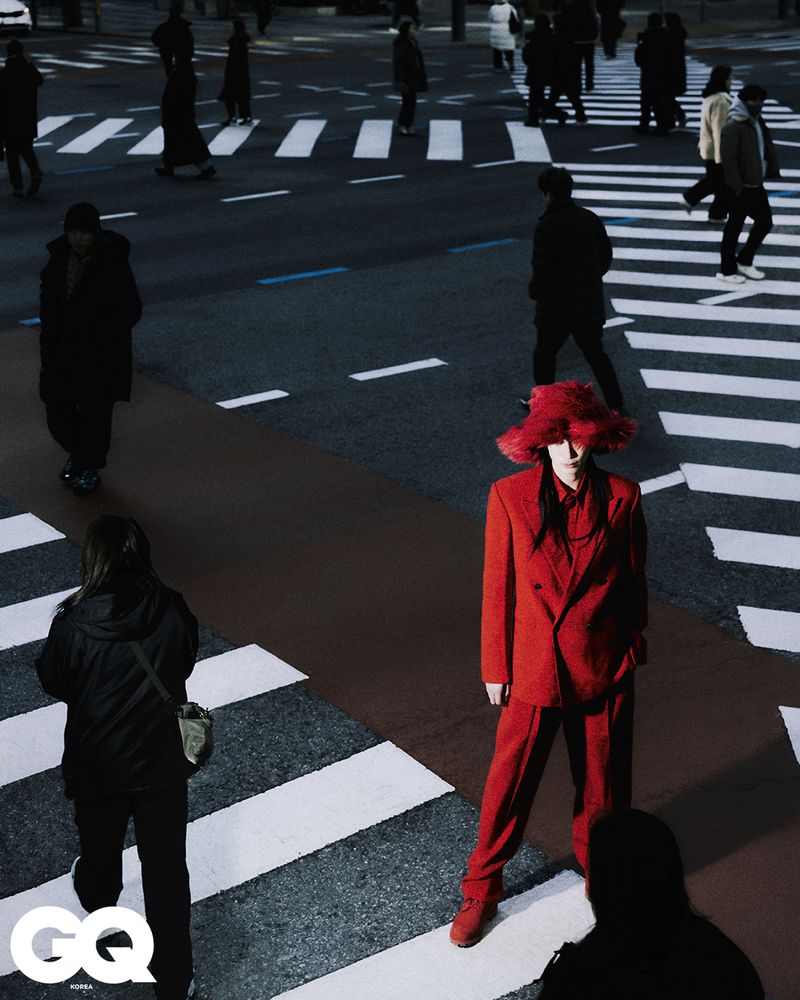 A fashion photo featuring a person in a striking red suit and hat amidst pedestrians crossing a city street with crosswalks and road markings.