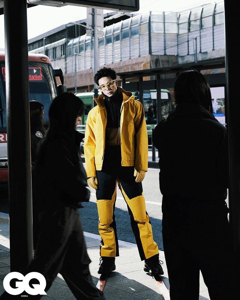 Man in yellow jacket and pants standing at a bus stop surrounded by blurred figures of people in an urban environment with a bus nearby.