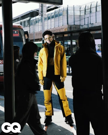 Man in yellow jacket and pants standing at a bus stop surrounded by blurred figures of people in an urban environment with a bus nearby.