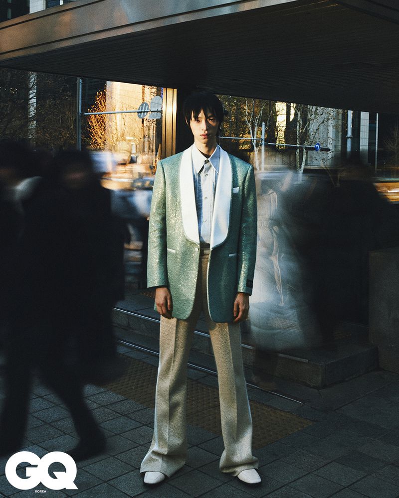 Fashion editorial shot of a young adult male in a shiny green blazer standing in an urban environment with blurred figures. 