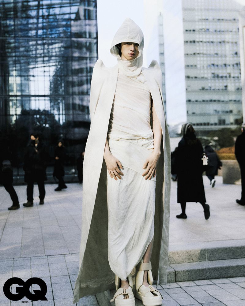 A person wearing a hooded white avant-garde outfit with platform shoes standing on a sidewalk against a city building backdrop.